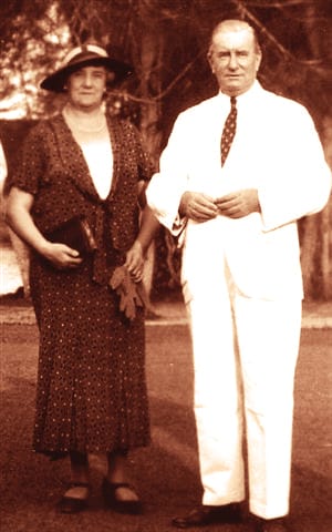 A man in a white suit and tie stands next to a woman in a dark polka dot dress holding a purse and hat.